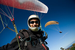 Paraglider wearing a helmet and sunglasses smiling at the camera, with colorful parachute lines and another paraglider in the background.
