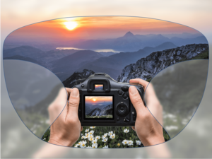 View through a pair of glasses showing hands holding a camera, capturing a mountain landscape with a sunset
