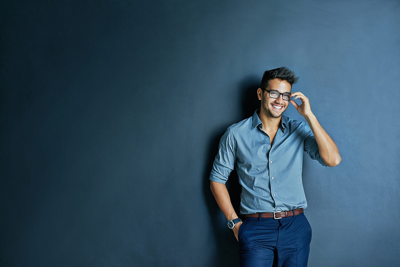 Smiling man in a blue shirt and trousers adjusting his glasses while leaning against a dark blue wall.