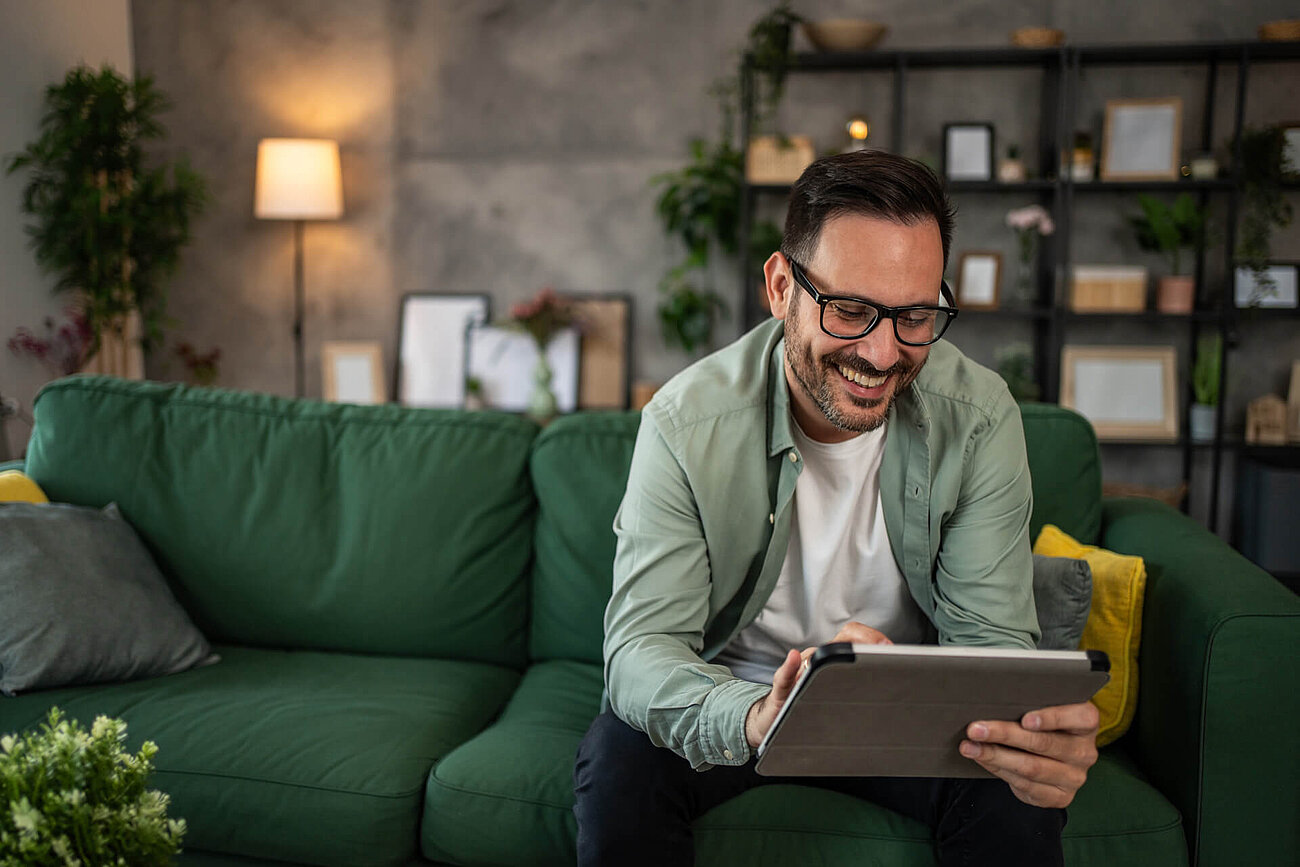 Smiling man with glasses sitting on a green sofa, using a tablet in a cozy living room.