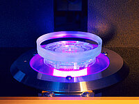 Glass lens being precisely processed or inspected in a machine under violet light.