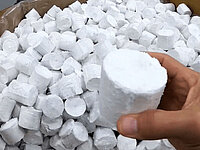 Hand holding a cylindrical white pellet above a container filled with many similar pellets.