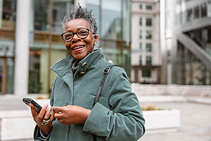 Smiling elderly woman in a green coat holding a smartphone, standing outdoors in an urban environment.