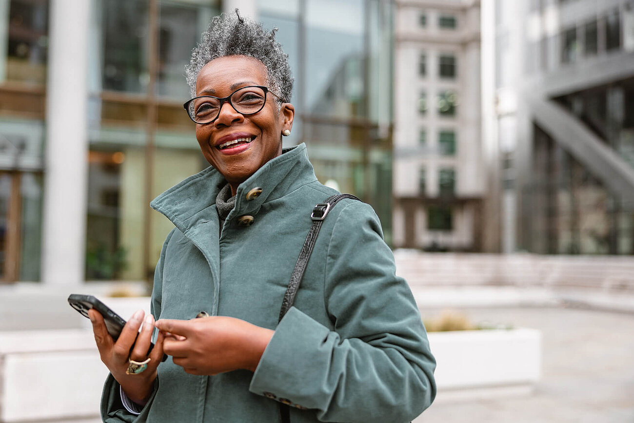Smiling elderly woman in a green coat holding a smartphone, standing outdoors in an urban environment.