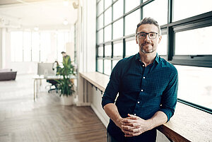 Smiling man with glasses standing by a large window in a bright, modern office space.