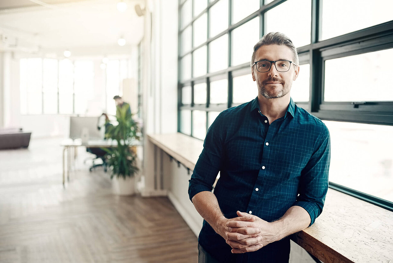 Smiling man with glasses standing by a large window in a bright, modern office space.