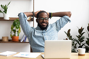 Smiling man with glasses sitting at a desk with a laptop, stretching with his hands behind his head, with plants in the background.