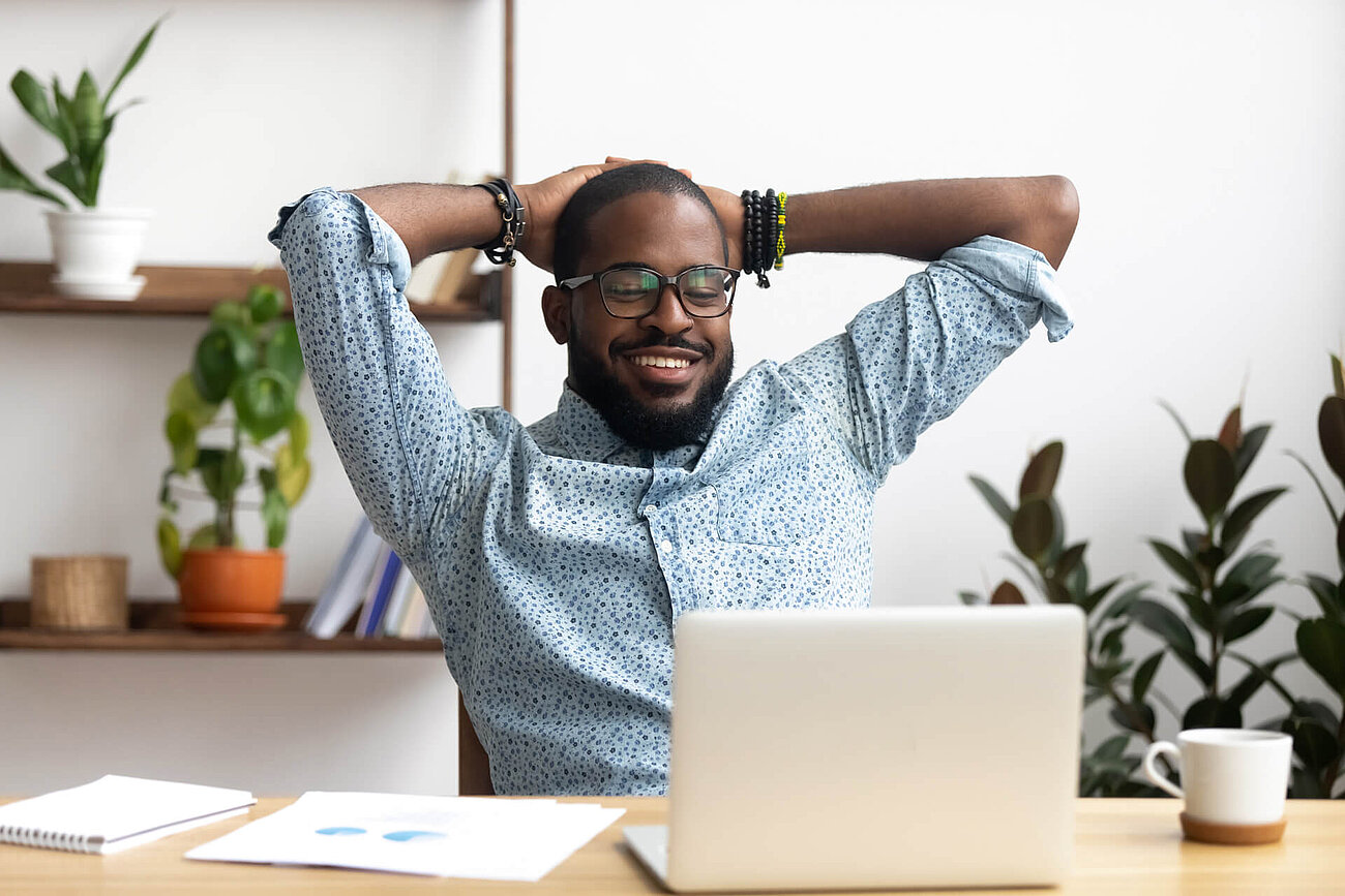 Smiling man with glasses sitting at a desk with a laptop, stretching with his hands behind his head, with plants in the background.