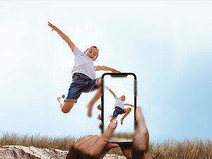 Child jumping joyfully in the air while being photographed with a smartphone in the foreground.