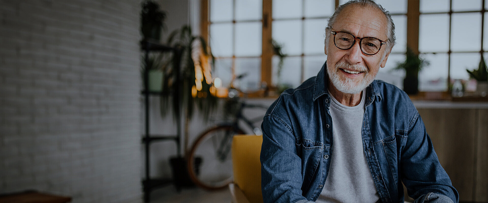 Great comfort, strong visual fields Smiling elderly man with glasses and a beard, sitting indoors in a bright room with plants and a bicycle in the background.