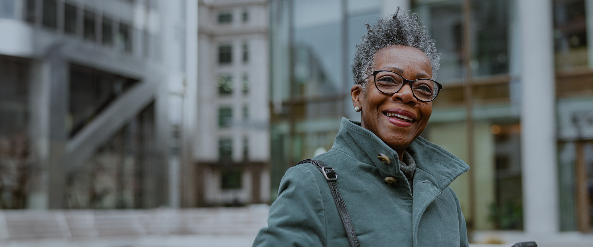 Smiling elderly woman wearing a coat and glasses, standing outdoors in an urban setting.