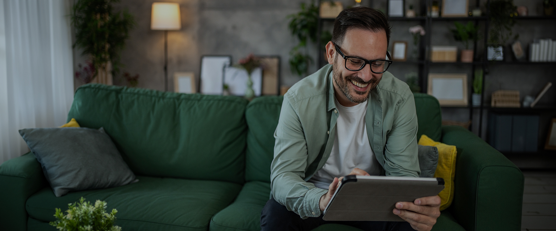 Smiling man with glasses sitting on a green sofa, using a tablet in a cozy living room.