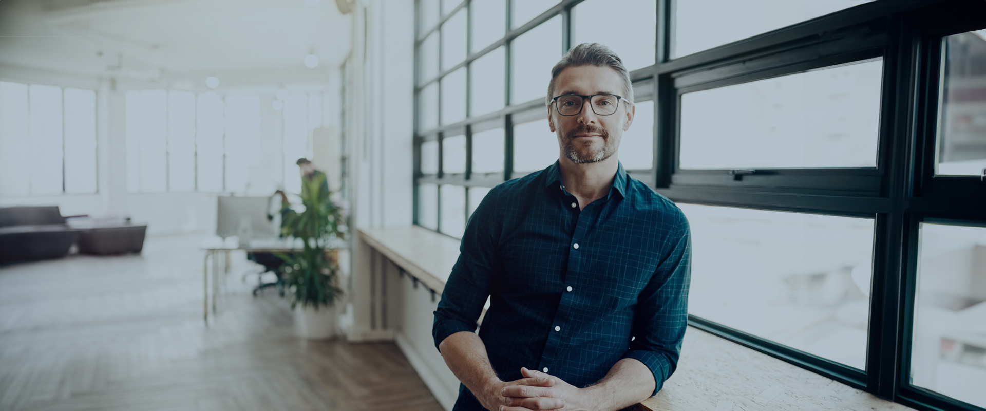 Man with glasses and a beard sitting by a large window in a bright, modern office space.