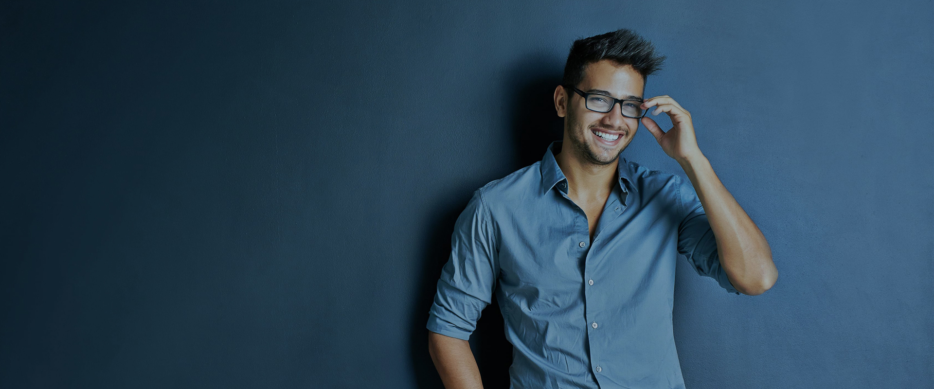 Smiling man in a blue shirt adjusting his glasses while standing against a dark blue wall.