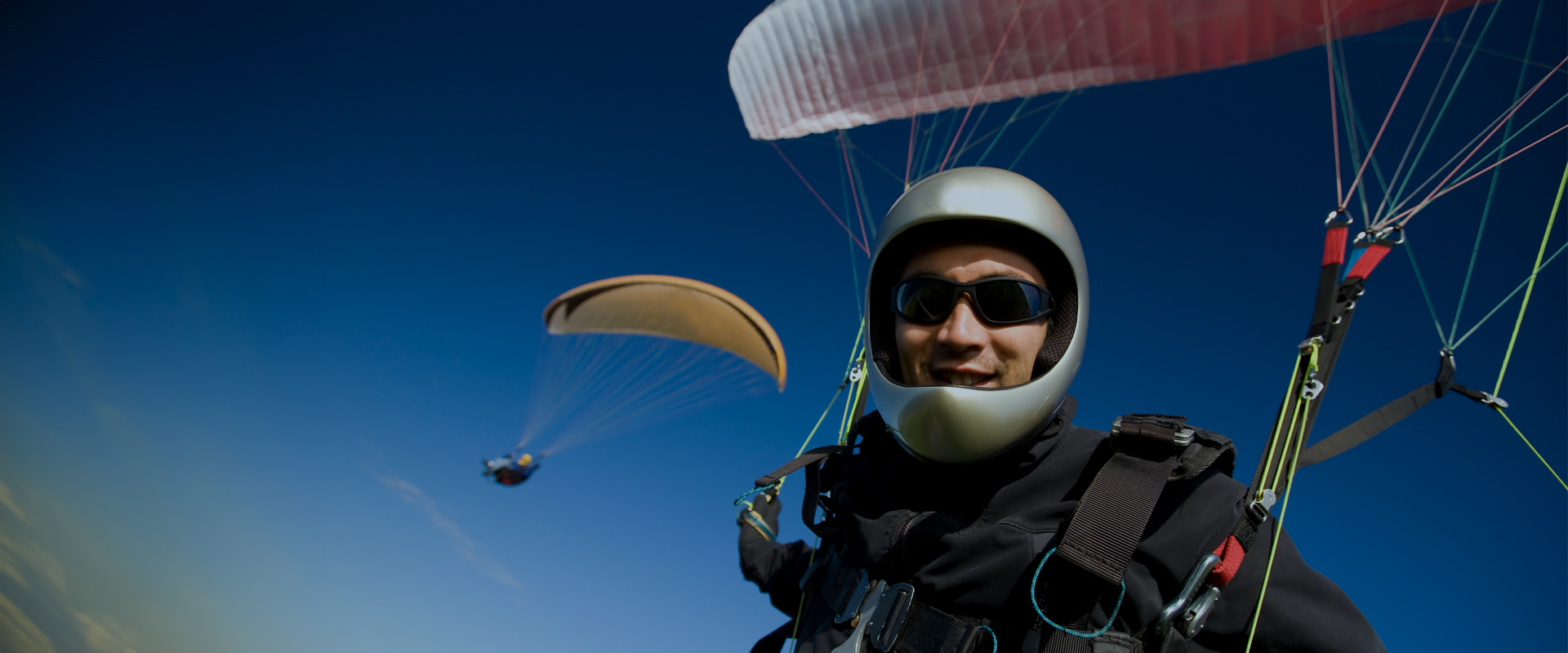 Paraglider wearing a helmet and sunglasses smiling at the camera, with another paraglider visible in the background against a clear blue sky.