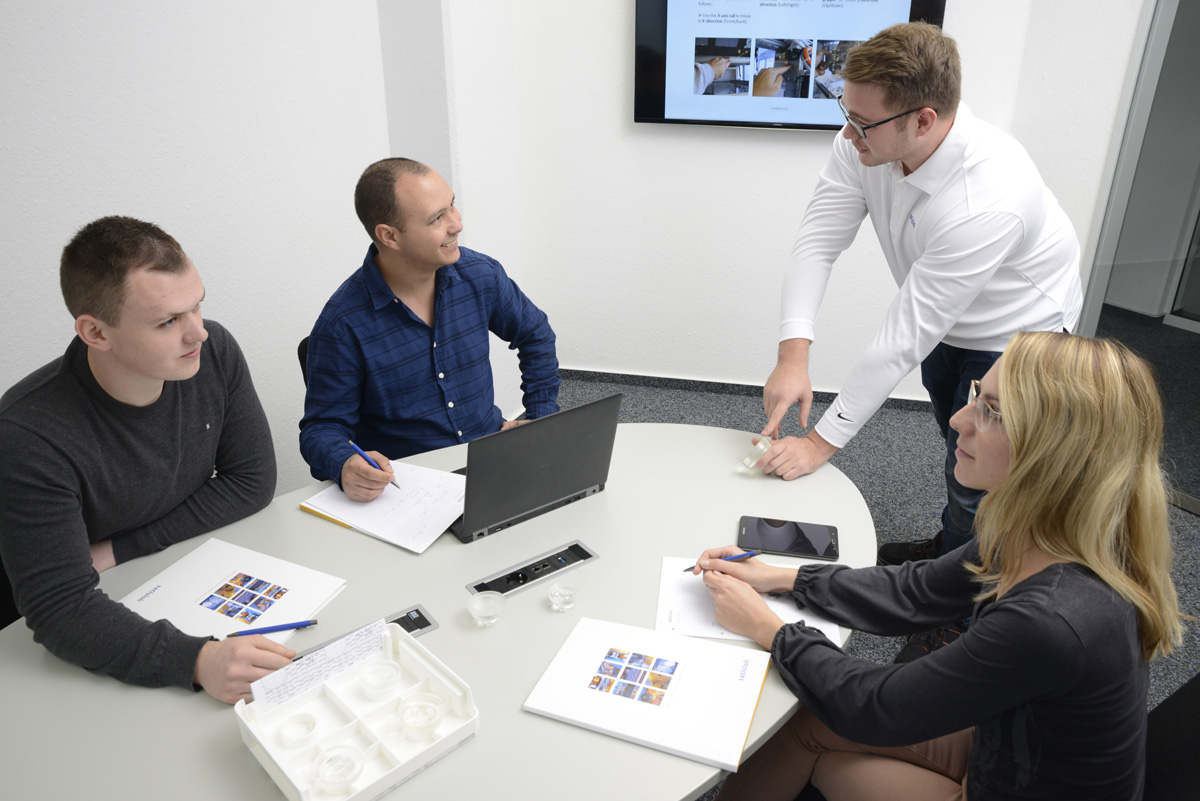 Four people in a meeting room discussing around a table with laptops and documents.