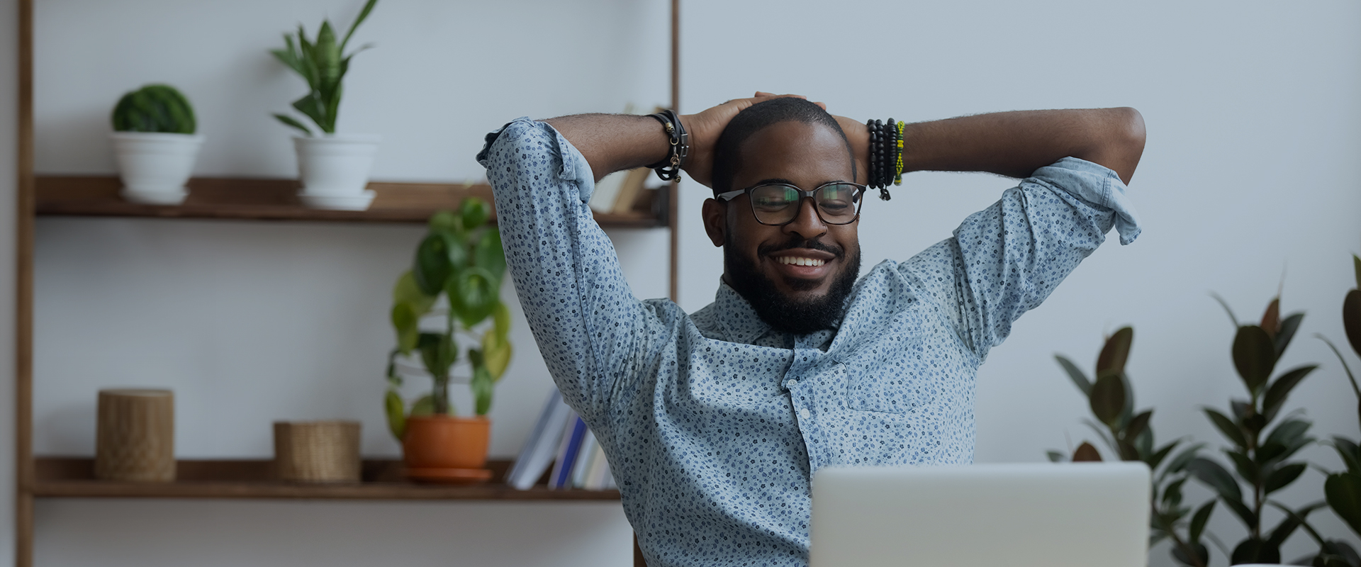 Wider view, better posture and superior comfort Smiling man with glasses sitting at a desk, stretching with his hands behind his head, with plants on shelves in the background